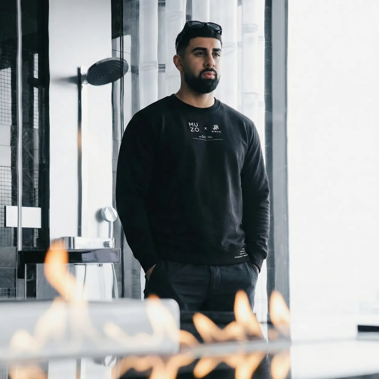 A man in the black MUZO x JR Strain sweatshirt standing by a window with his hands in his pockets, a fire trough burning in the foreground