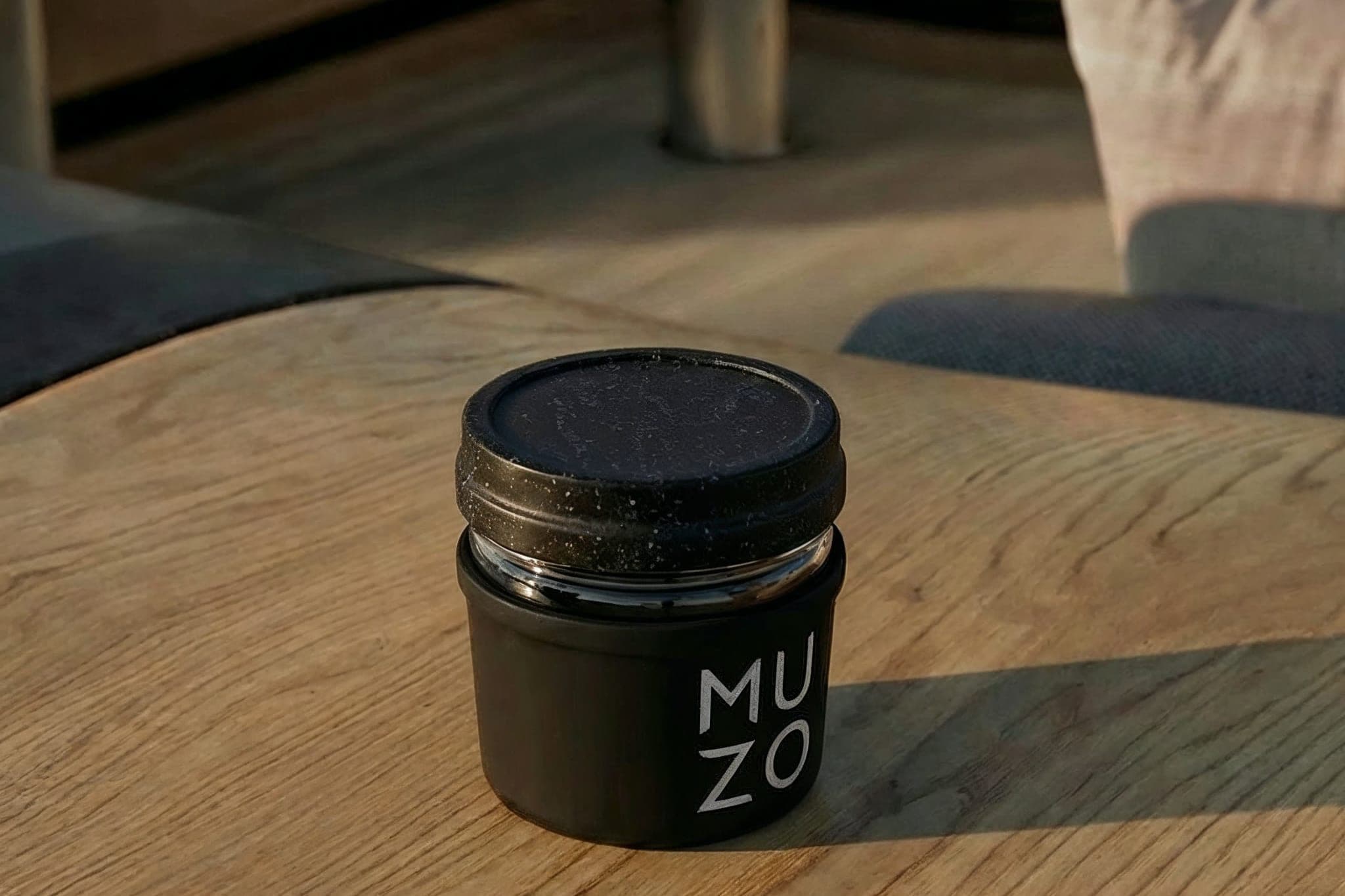 The matte black MUZO jar with its metal collar and the wordmark on the face, standing closed on a wooden table