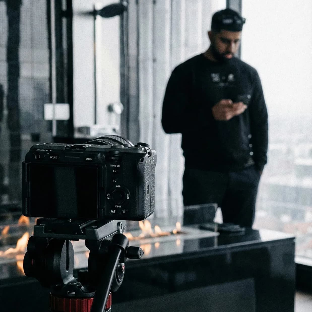 A mirrorless camera on a tripod in the foreground, and behind it, out of focus, the man in the MUZO sweatshirt and cap looking at his phone by the fire trough