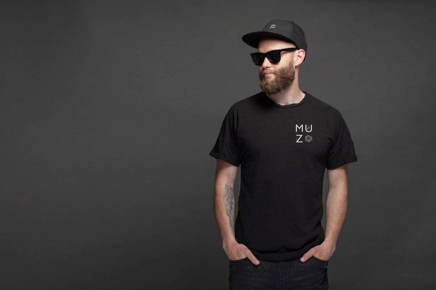 A bearded man in sunglasses, a black MUZO cap and a black MUZO tee, hands in his pockets, against a grey studio ground