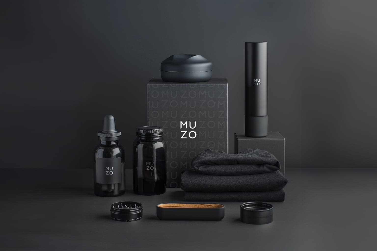 The MUZO welcome pack on grey: the box printed with the monogram field, a dropper bottle and a jar, a black vaporiser, a folded sweatshirt, a grinder, a wooden tray and a tin lid