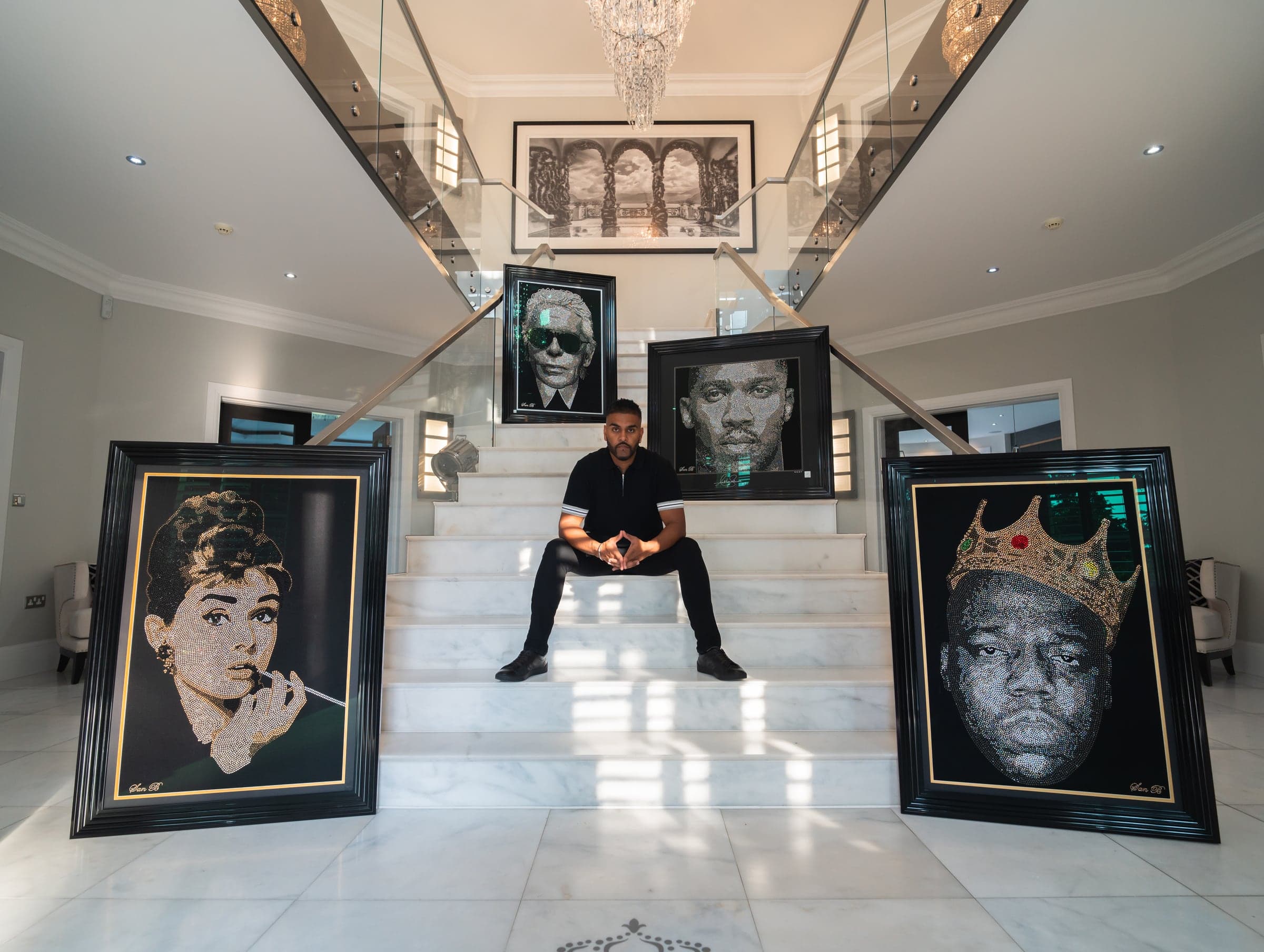 San B seated on a marble staircase, the Audrey Hepburn piece to his left and The Notorious B.I.G. to his right, the Karl Lagerfeld piece on the landing behind