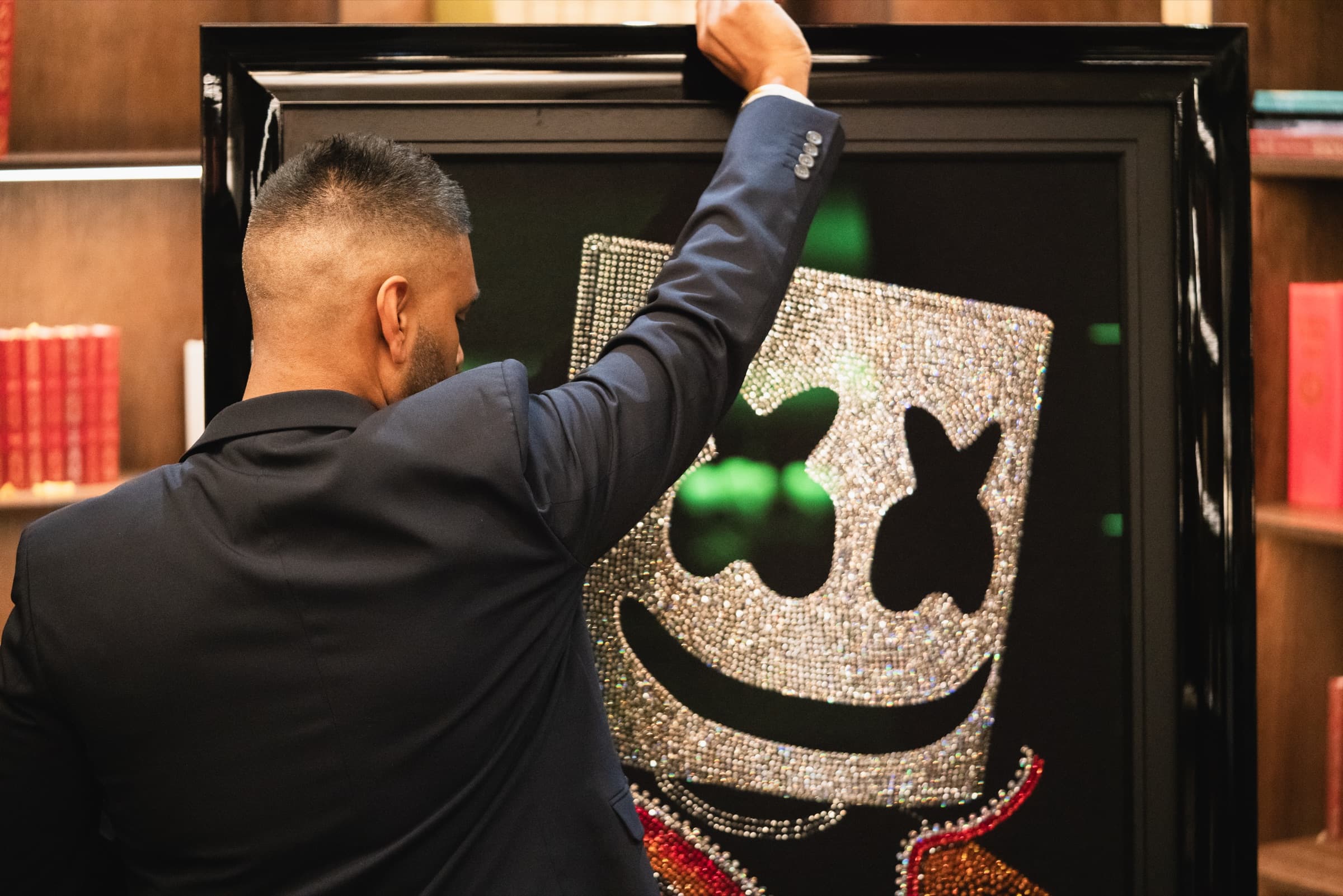 San B from behind, reaching up to set the framed Marshmello piece into the lit bookshelves of the panelled library