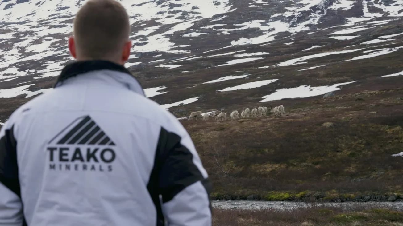 A man in a Teako jacket on the moor at the snow line, reindeer beyond him