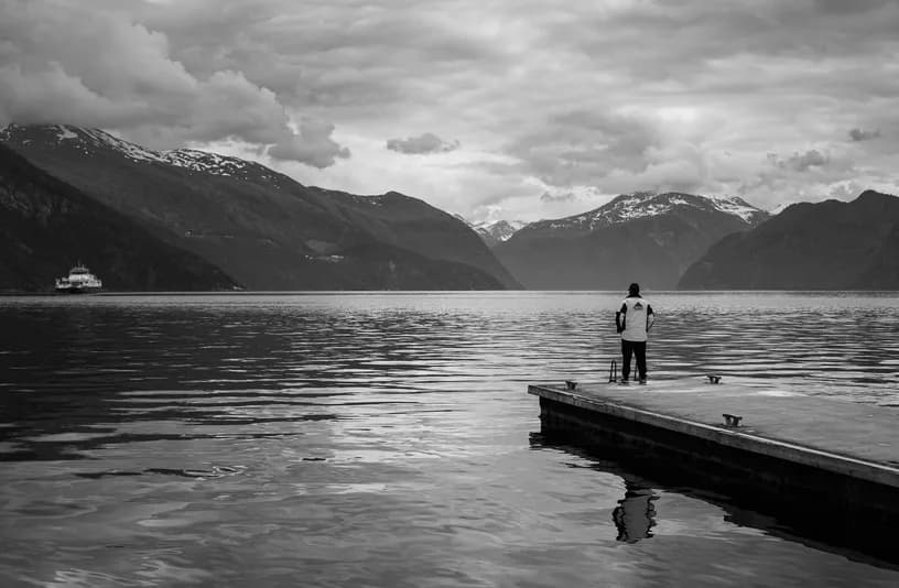 A man alone at the end of the jetty, the fjord and the mountains before him, in black and white