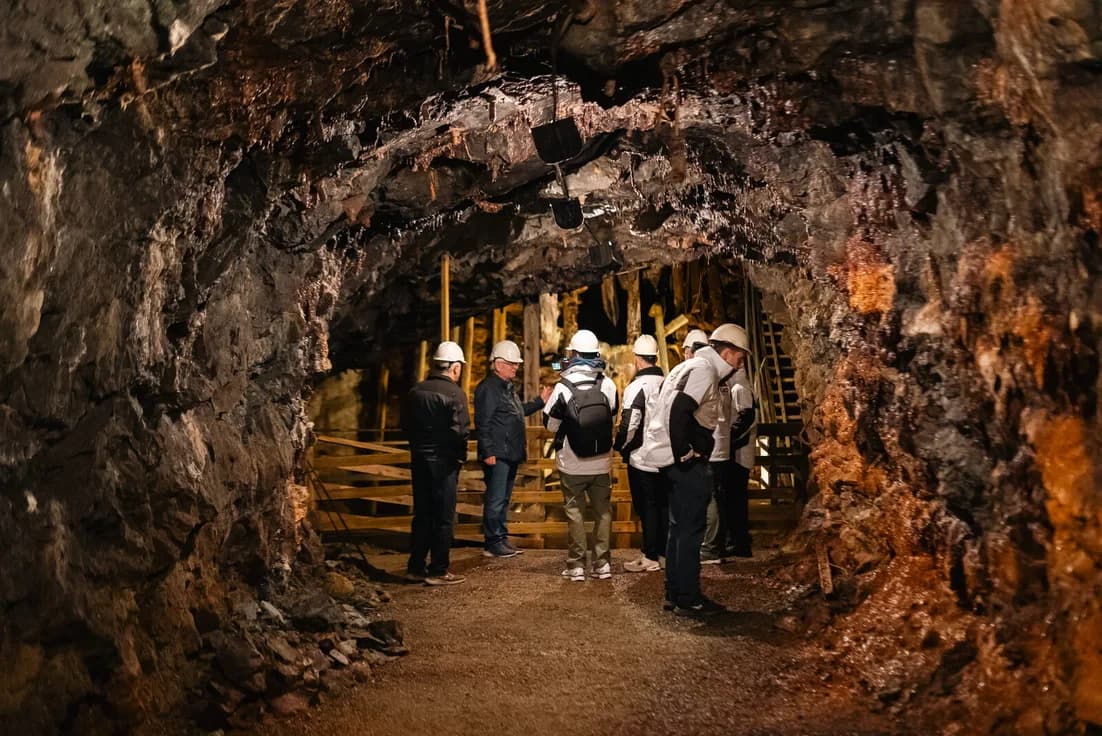 A group in hard hats walking out of a lit adit, the rock ochre and wet on both sides.
