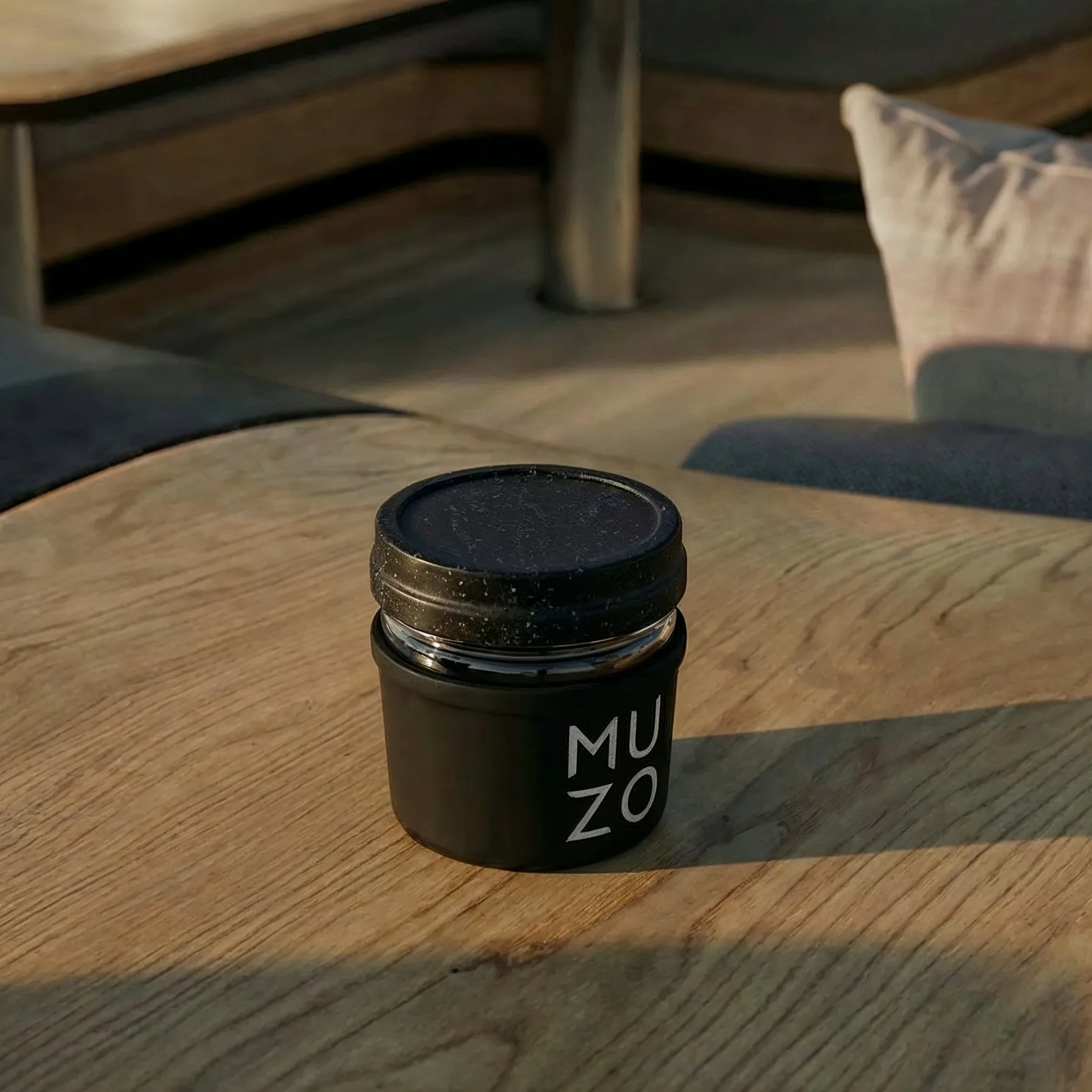 The matte black MUZO jar with its metal collar and the wordmark on the face, standing closed on a wooden table